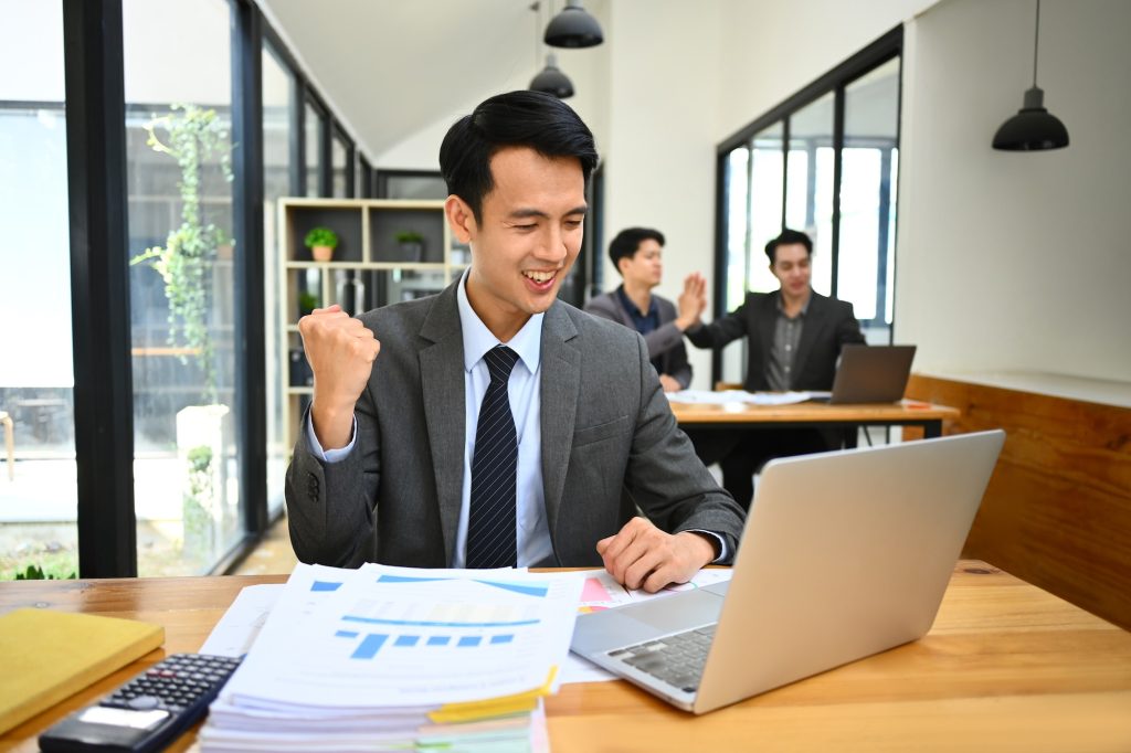 Excited asian male office worker looking at laptop screen, celebrating business success.