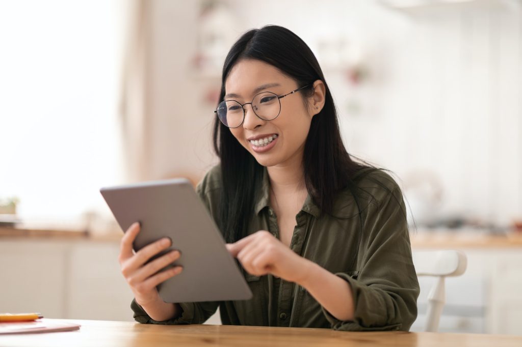 Positive young asian woman using digital tablet at kitchen