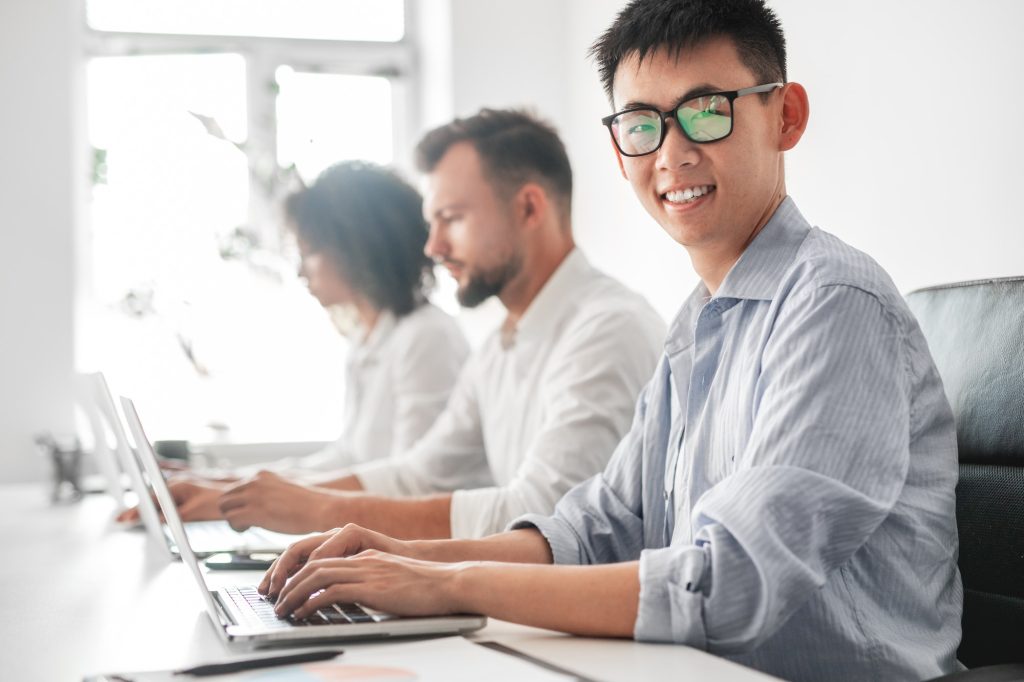 Smiling Asian male employee working on laptop near diverse colleagues in office